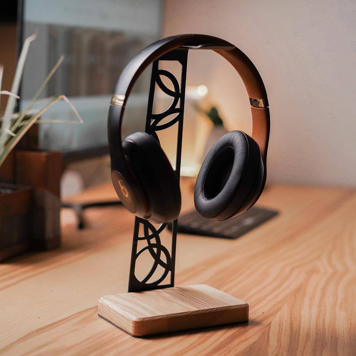 Handcrafted headphone stand with a smooth birch wood base and a black metal frame featuring intricate cutout patterns, holding over-ear headphones on a wooden desk in a modern workspace.